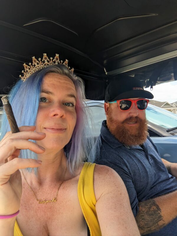 selfie of a person with blue hair wearing a tiara and holding a cigar with a man with a stylish beard looking on from behind