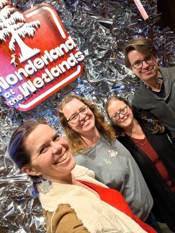 four people take a group selfie in front of a sparkly wall with a sign for Wonderland at the Wetlands