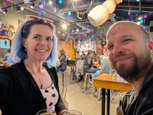 a selfie of two people at a brewery decorated with an excessive amount of holiday string lights