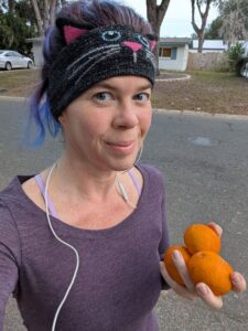 selfie of a woman with a headband probably out running and also holding three small oranges