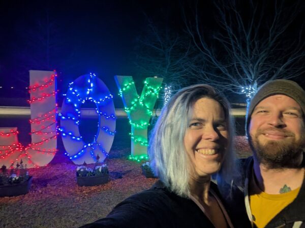 two people taking a selfie in a night time park with three large letters covered in lights spelling JOY in the background