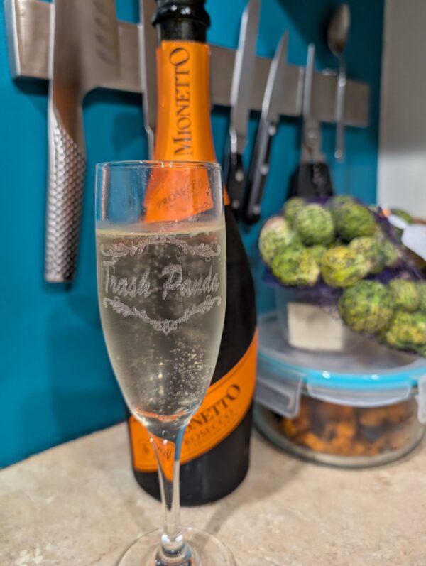 a champagne flute engraved with the words Trash Panda is full of bubbly and blurry in the background is a bottle and some cooking ingredients with a knife rack beyond them